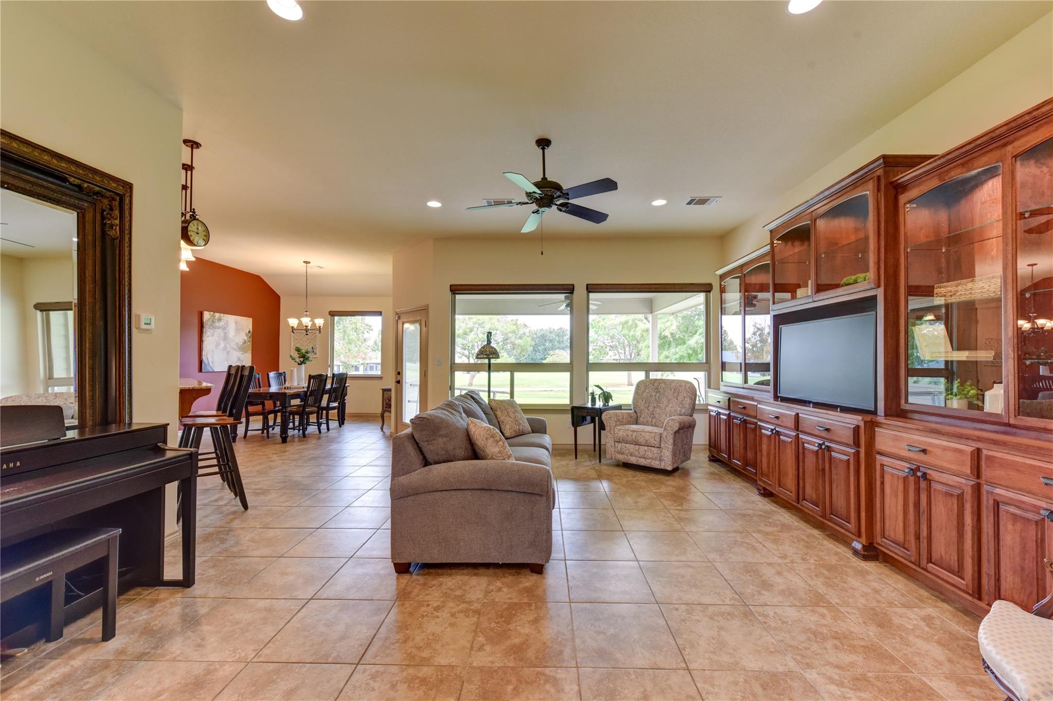 Open-plan living and dining room with built-in wood cabinetry, a piano, and wide windows overlooking a lawn.