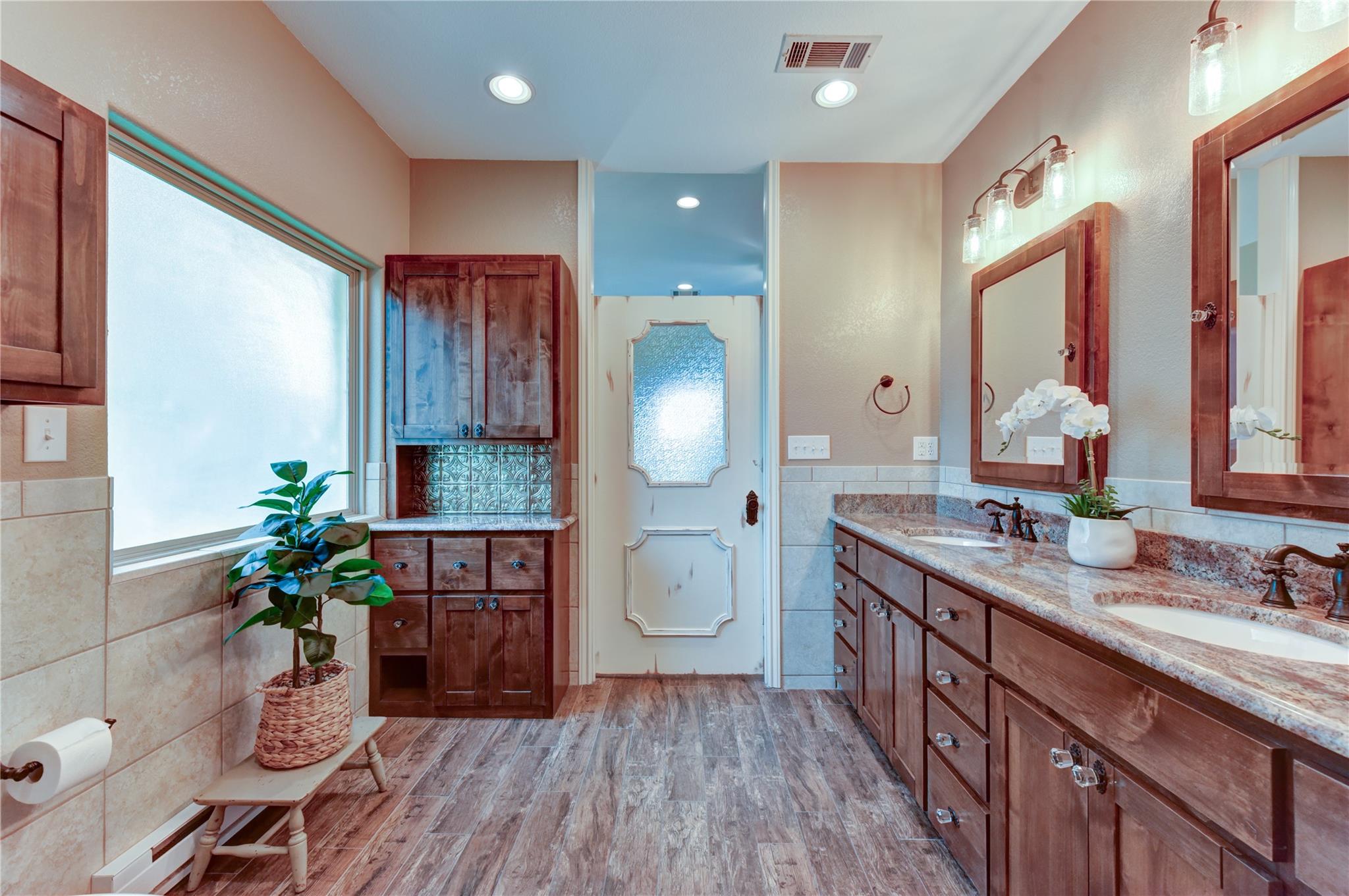 Bathroom with a long double-sink wood vanity, stone counters, framed mirrors, and a frosted-glass door.