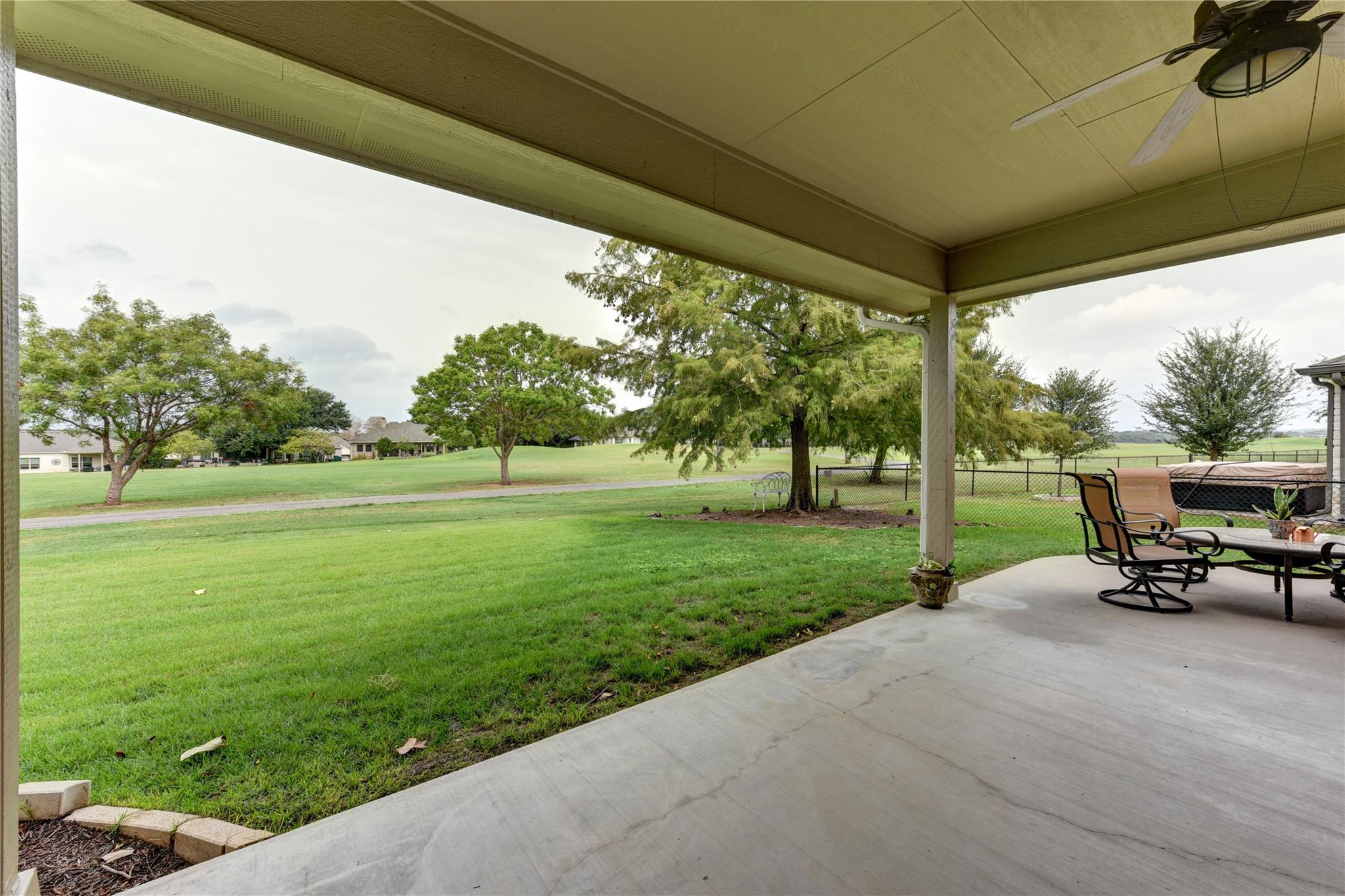 Covered patio overlooking a broad green lawn and mature trees, with outdoor seating and a fenced hot tub.