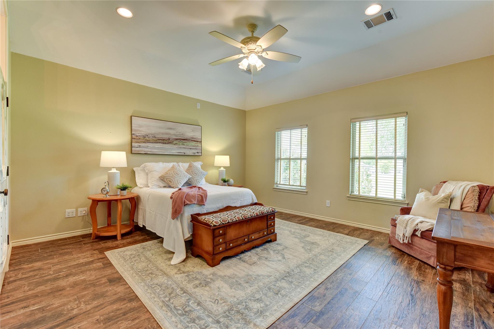 Furnished bedroom with a white bed, wooden storage bench, two windows, and a ceiling fan.