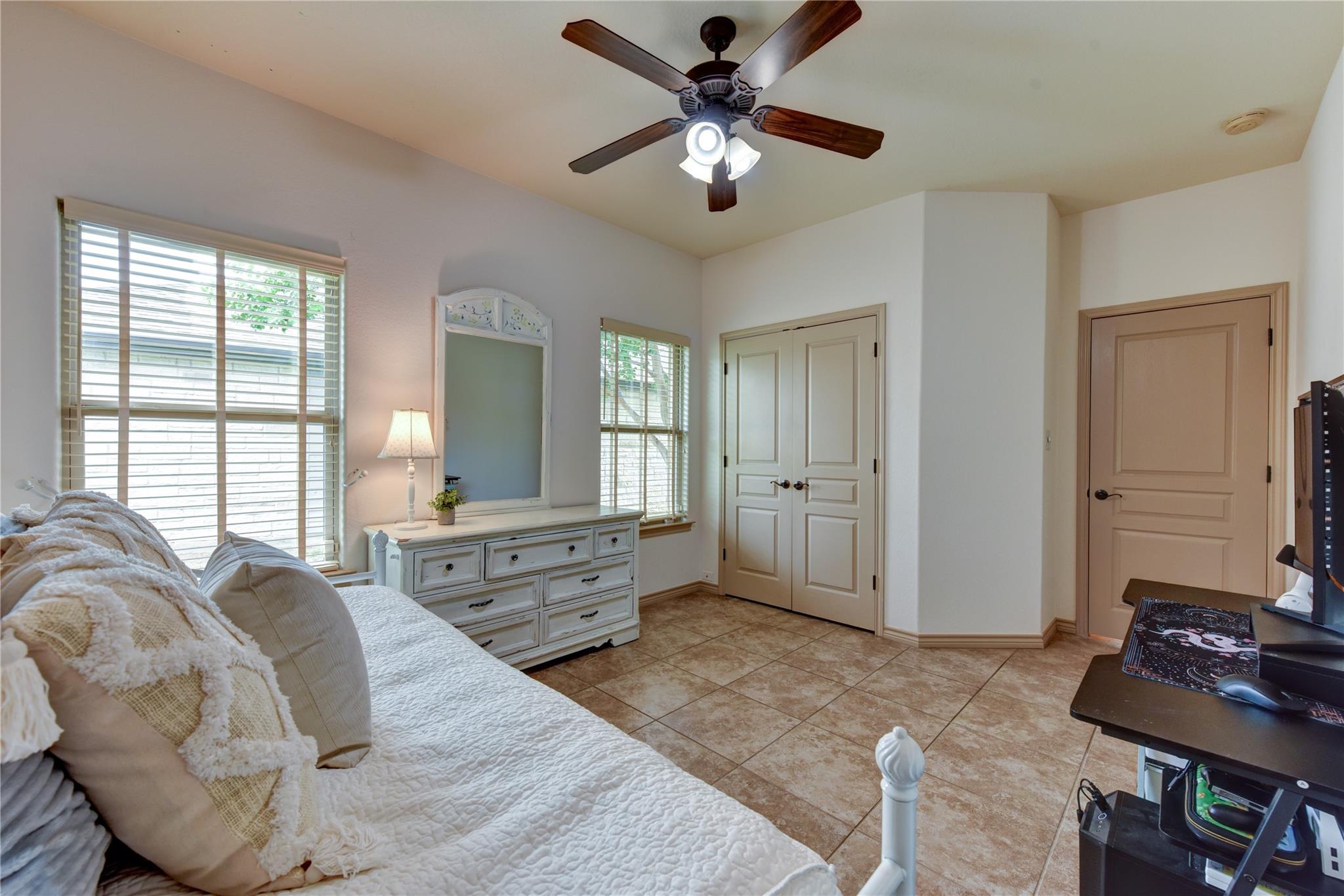 Furnished bedroom with a white bed, dresser between two windows, tiled floor, and ceiling fan.