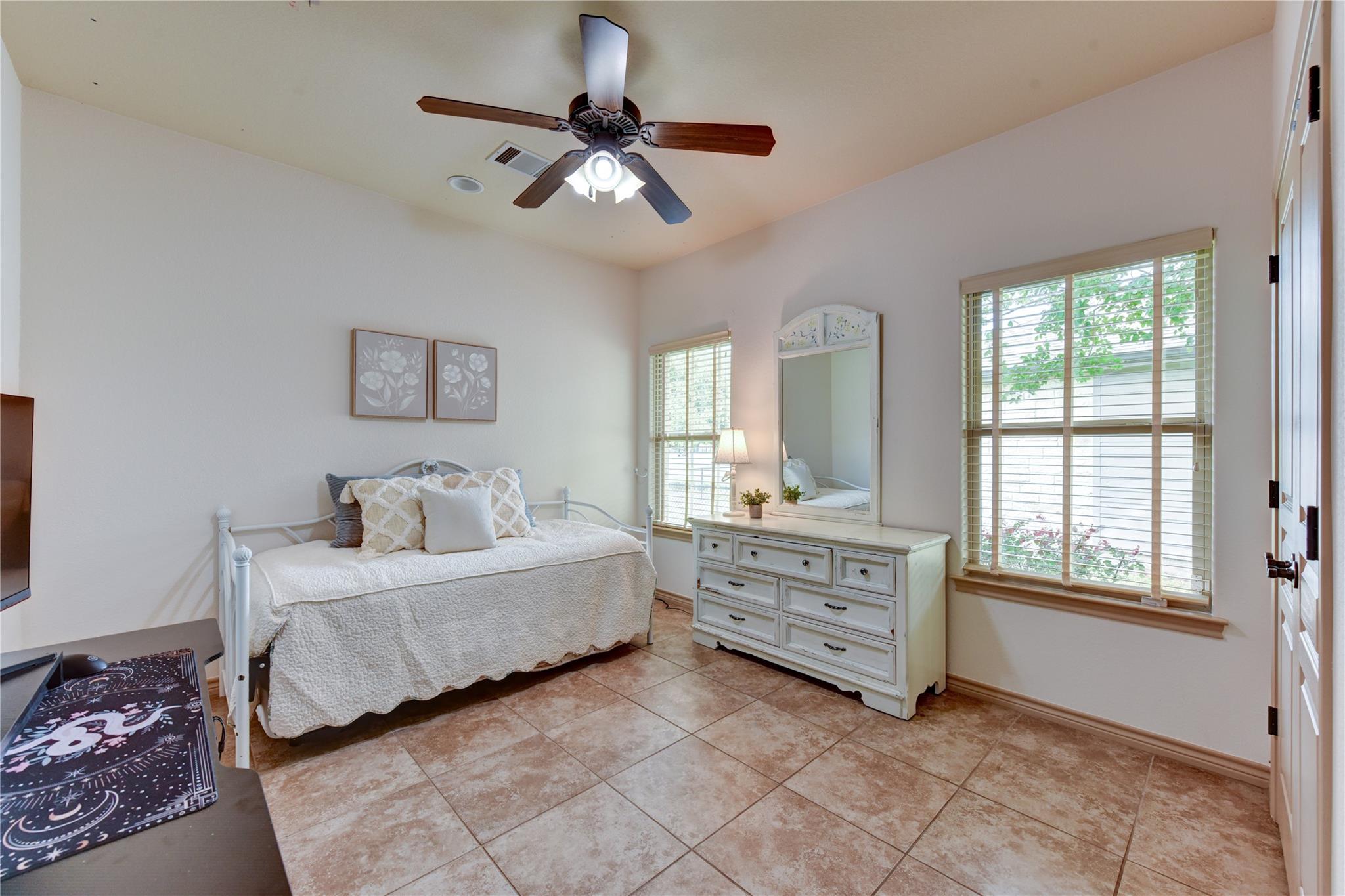 Furnished bedroom with a white bed, matching dresser and mirror, and two windows with blinds.