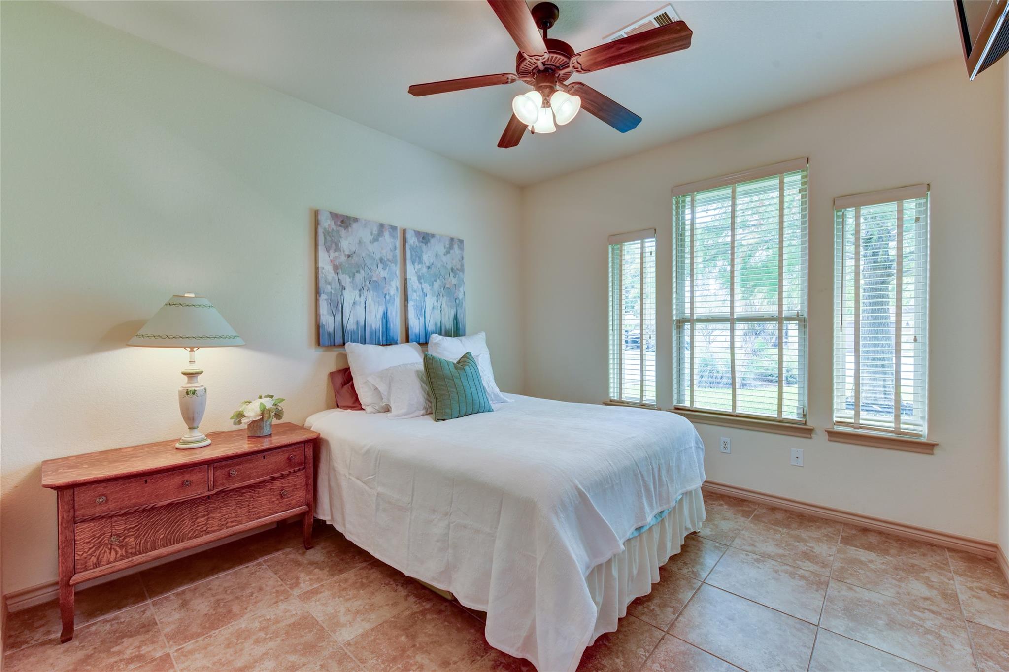 Furnished bedroom with a white bed, wood dresser, ceiling fan, and three bright windows.