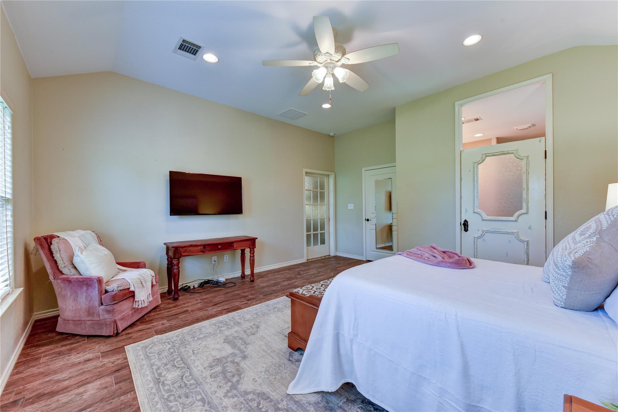 Bedroom with a large bed, ceiling fan, and pink chair beside a wall-mounted television.