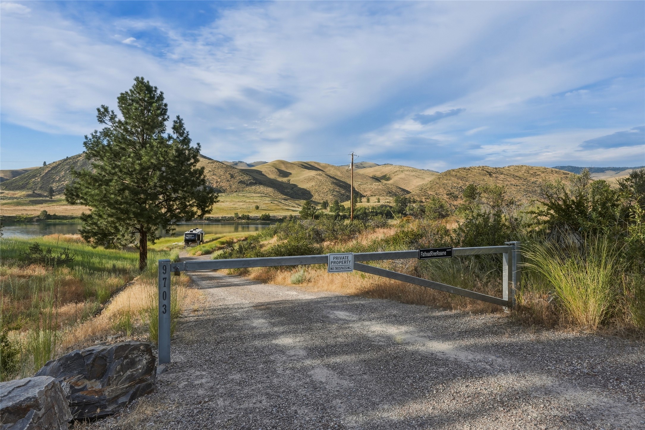 Gravel property entrance with a 9703 gate, river, pine tree, and rolling hills.