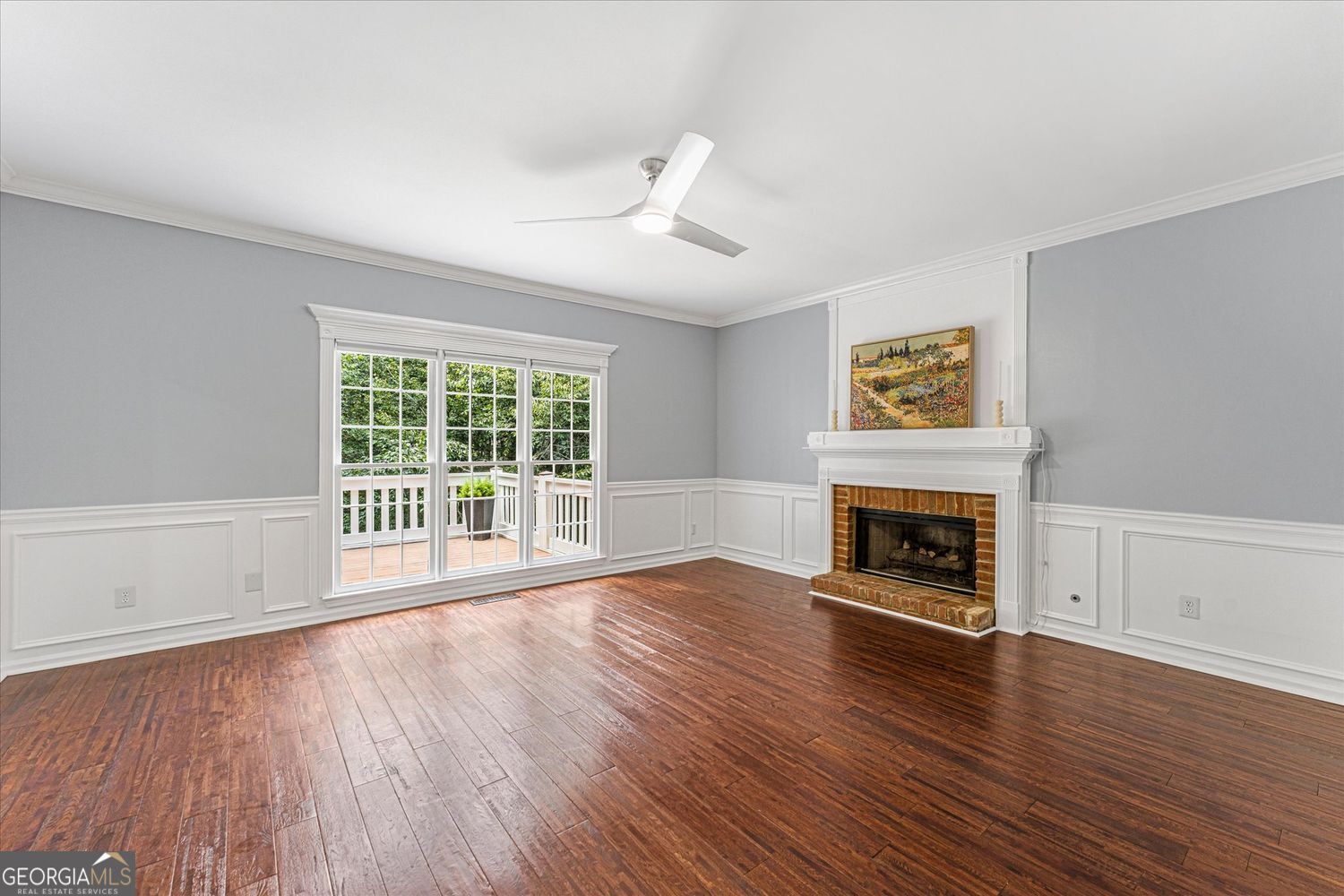 Empty living room with dark hardwood floors, a brick fireplace, and a window overlooking a deck.