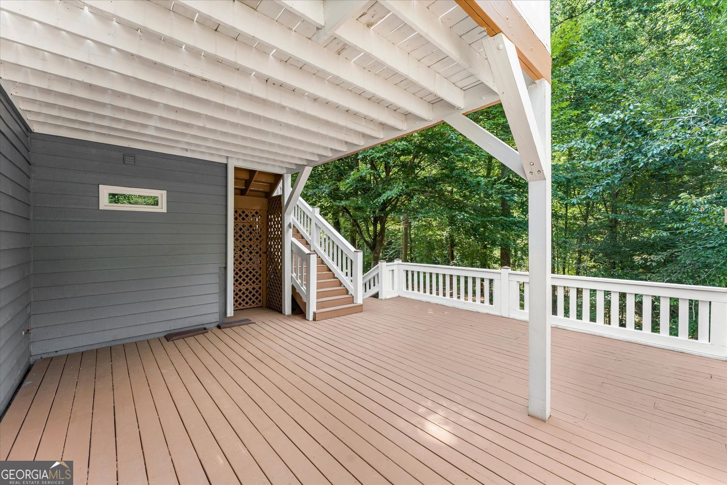 Covered wooden deck with white railings, stairs, and a view of surrounding trees.