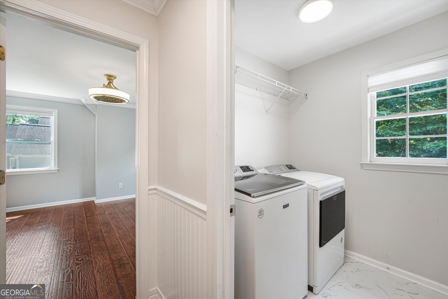 Laundry room with washer and dryer, wire shelf, and window overlooking trees.