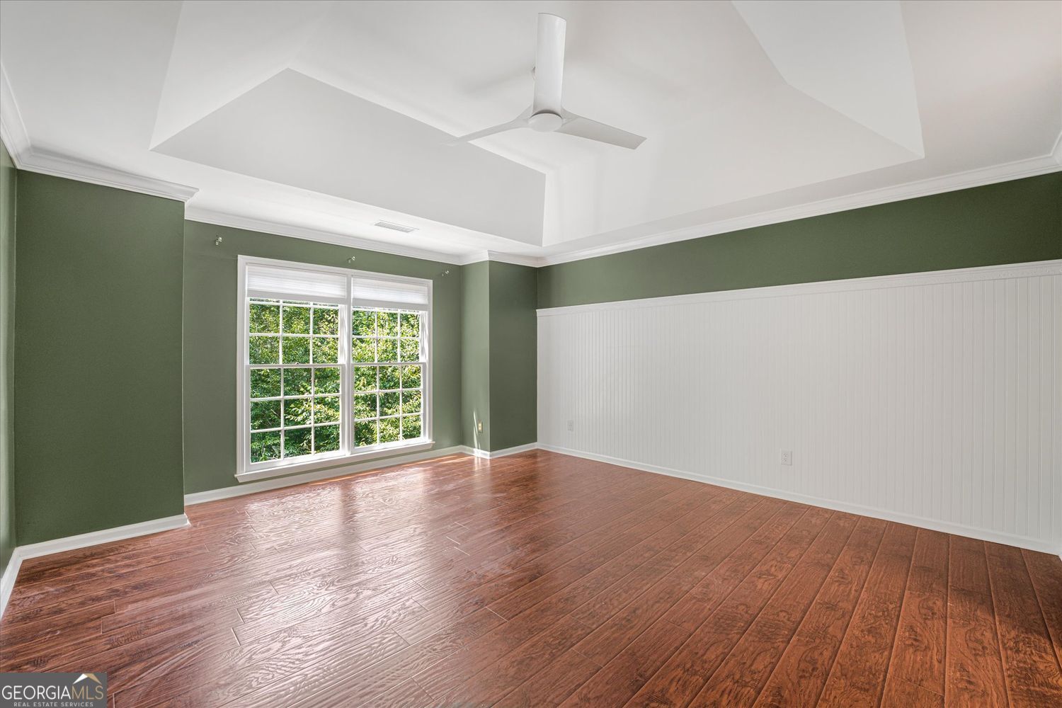 Empty room with reddish-brown wood floors, green walls, white beadboard, and a large window.