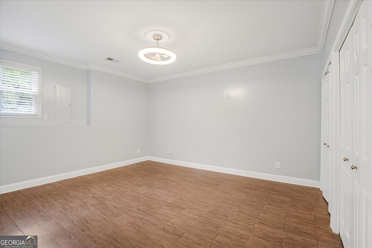 Empty bedroom with brown wood-look tile flooring, white closet doors, and a small window with blinds.