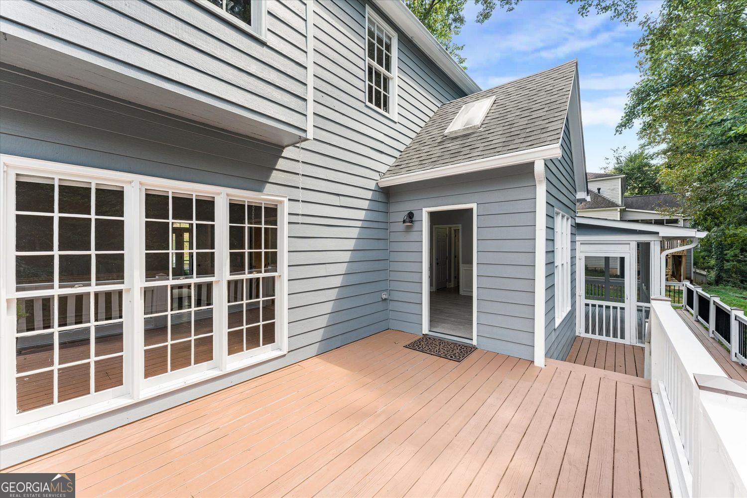 Rear deck with white railings beside a gray-sided house with large windows and an open doorway.
