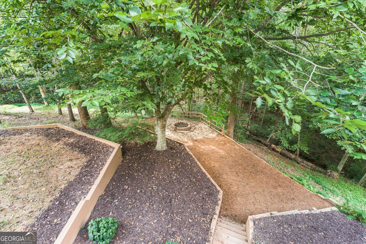 Wooded backyard with terraced garden beds, a pine-needle path, and a circular stone fire pit.