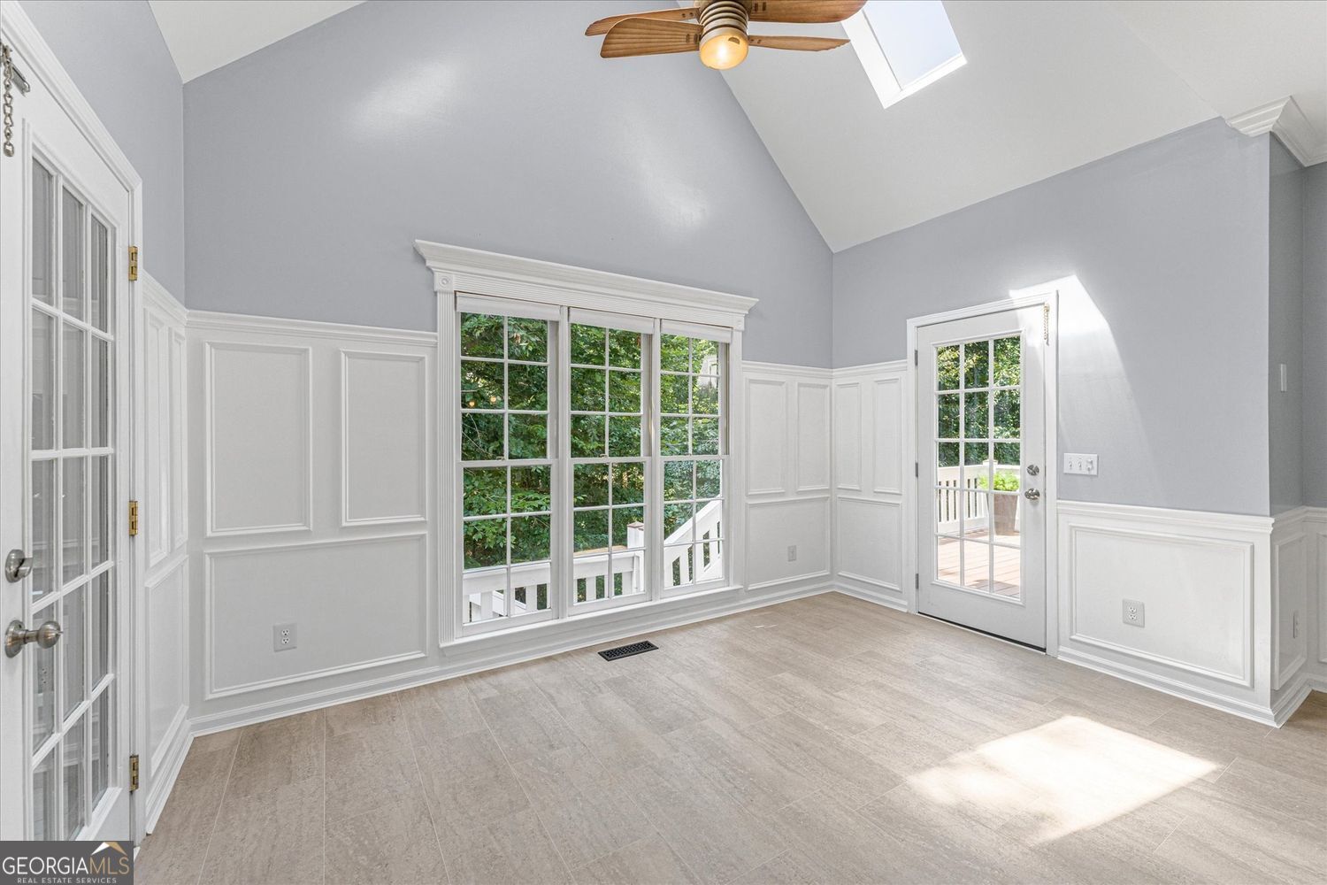 Bright vacant room with vaulted ceiling, white wainscoting, large windows, and glass doors to a deck.