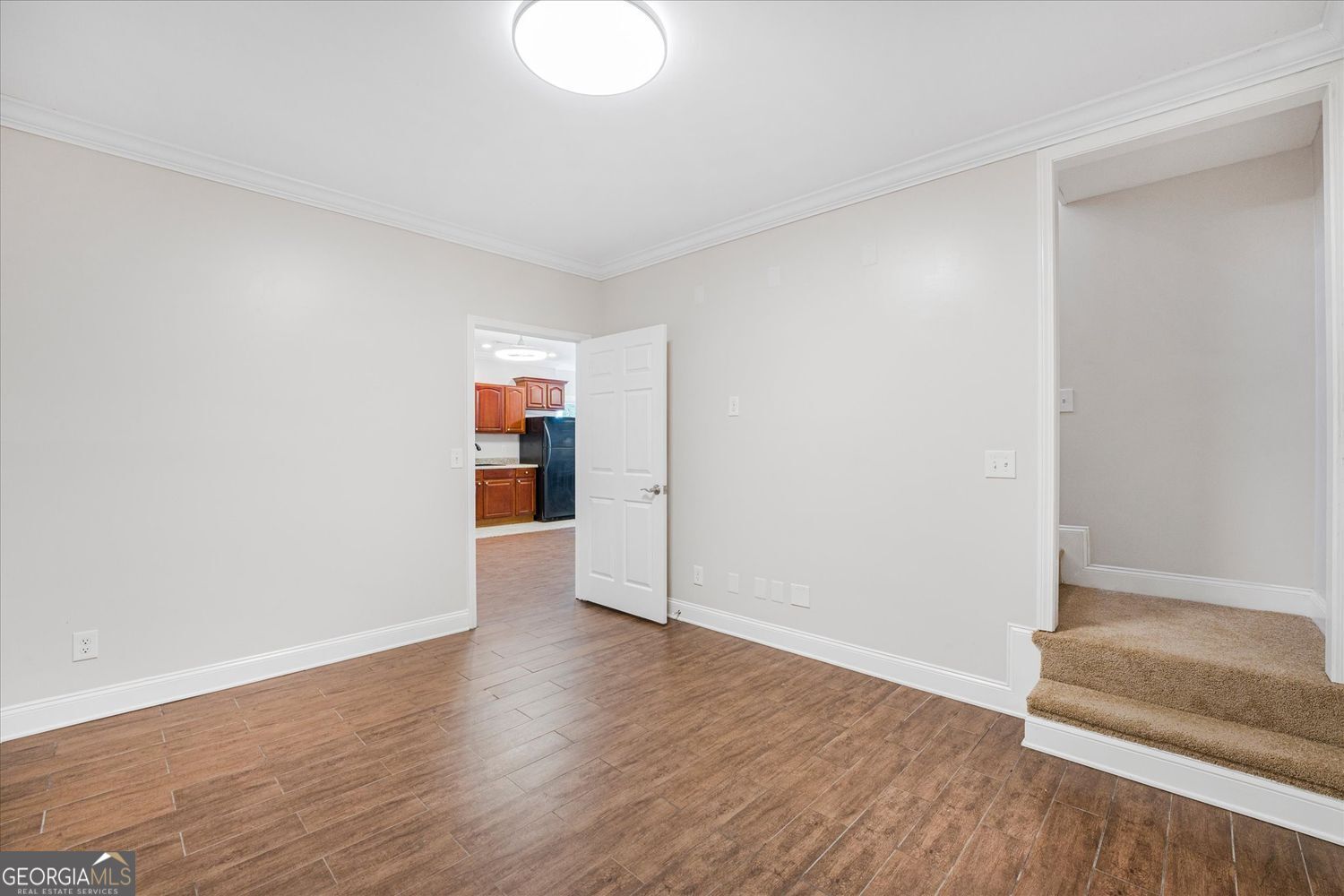 Empty room with wood-look tile flooring, opening to a kitchen and carpeted steps.