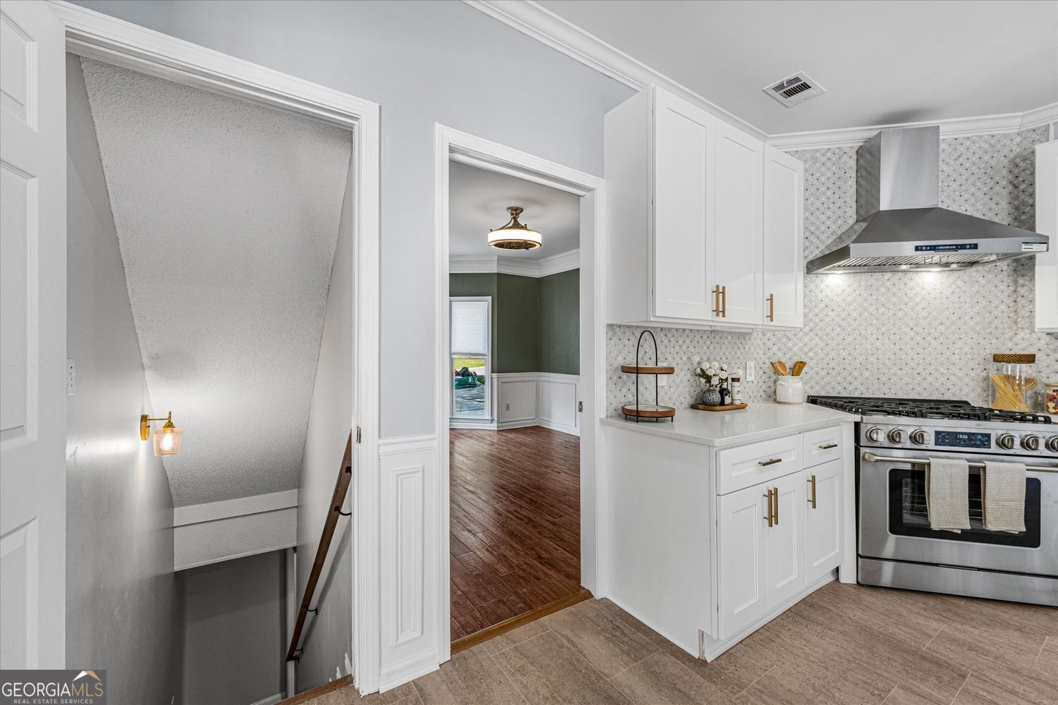 Kitchen with white cabinets, brass hardware, patterned backsplash, and stainless steel range beside an open staircase.