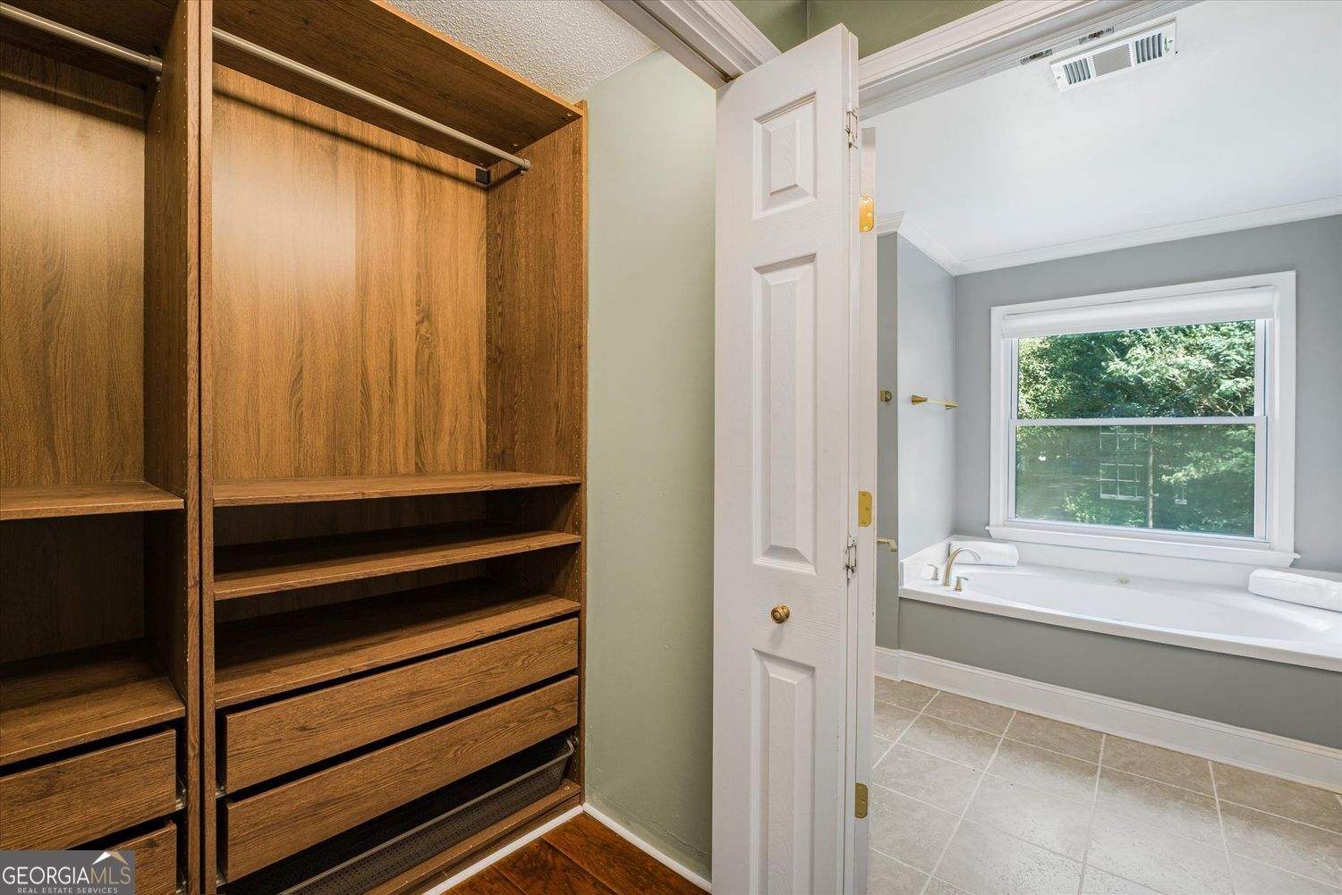 Walk-in closet with built-in wooden drawers beside a bathroom soaking tub.
