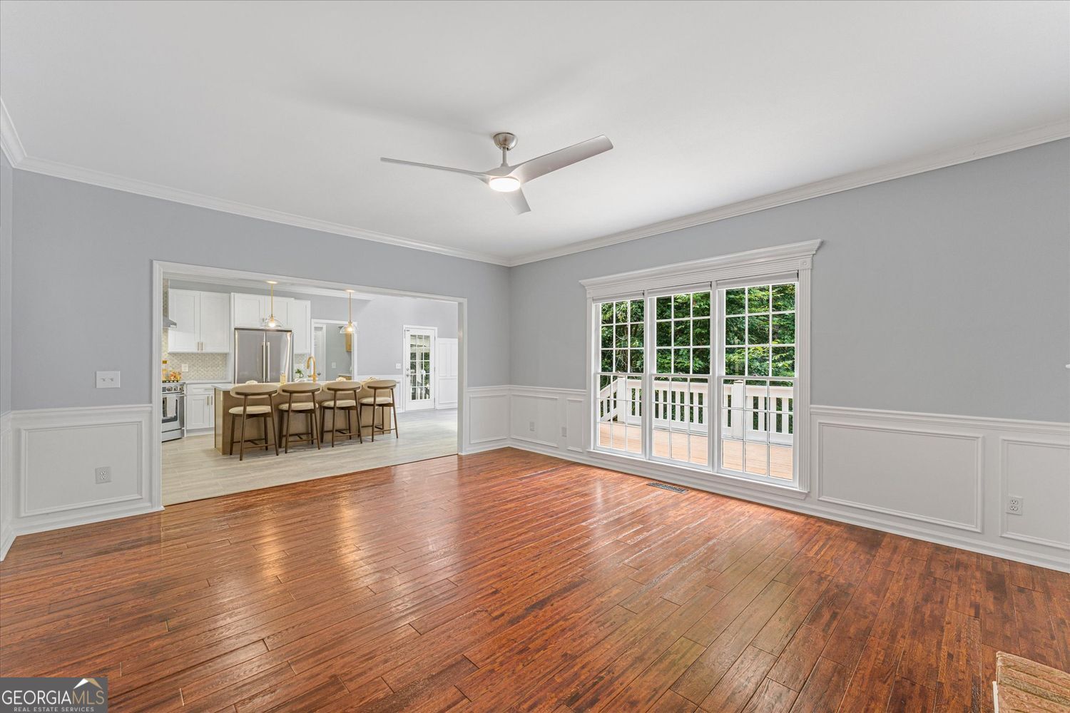 Empty living room with hardwood floors, white wainscoting, large windows, and an open view into the kitchen.