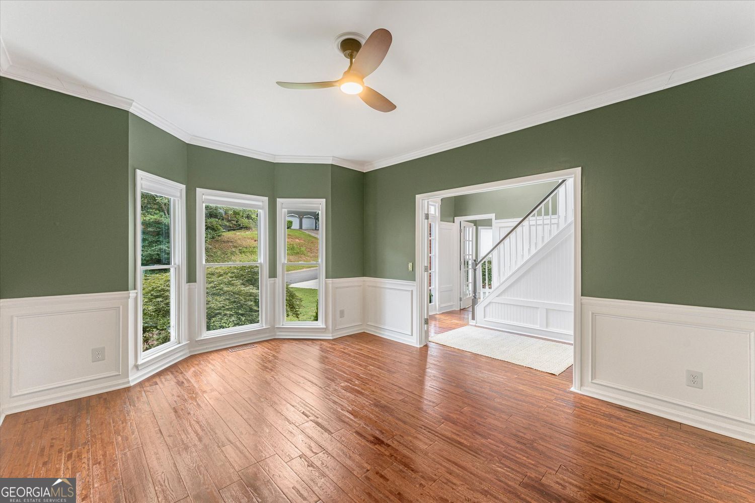 Empty room with green walls, white wainscoting, hardwood floors, three tall windows, and a view of the staircase.