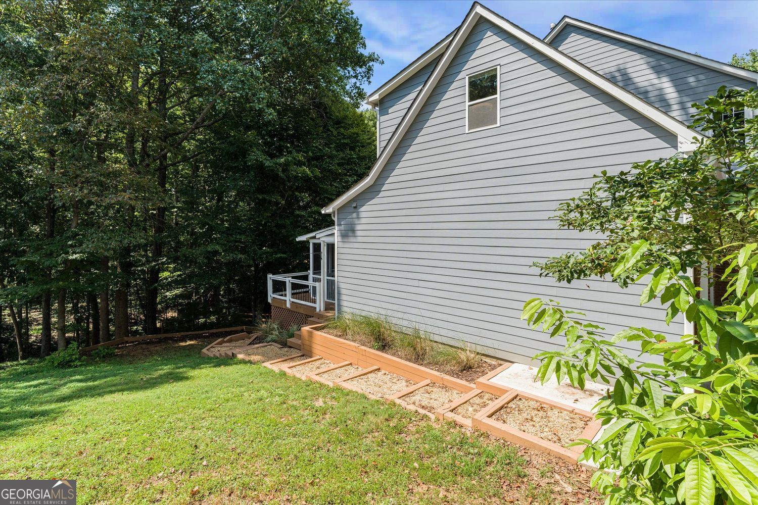 Gray two-story house beside a wooded yard, with a deck and gravel path through raised planting beds.