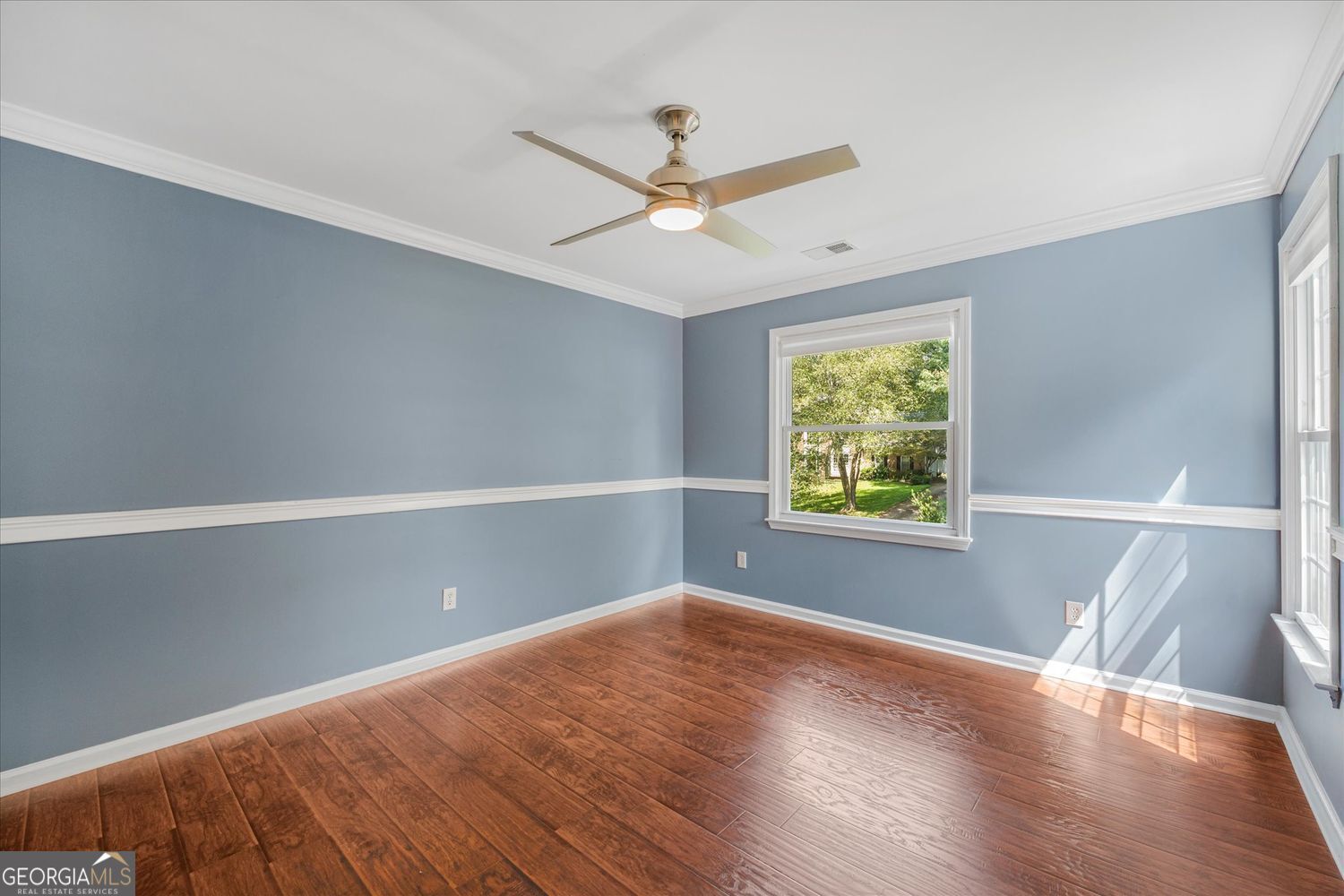 Vacant blue-walled room with wood-look floors, a ceiling fan, and windows overlooking a leafy yard.