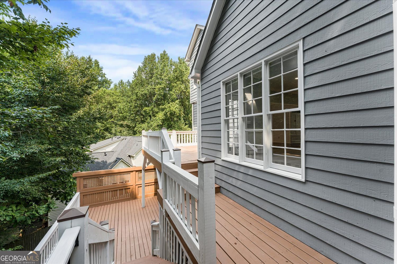 Multi-level wood deck with white railings beside gray siding and large windows, overlooking leafy trees.