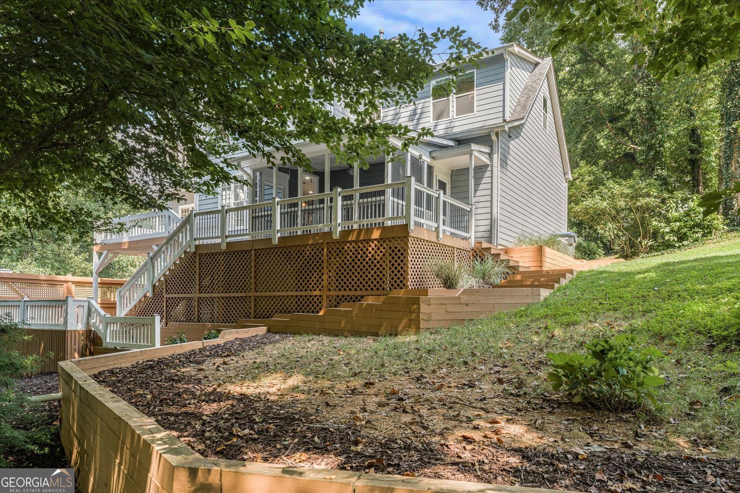 Gray two-story home with a raised deck and timber retaining walls on a wooded slope.
