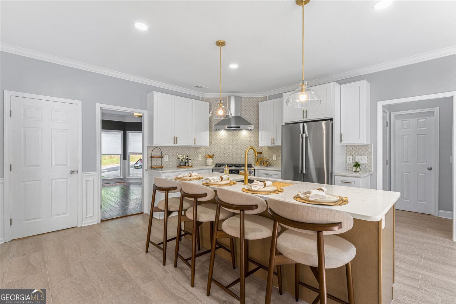 Staged kitchen with a large island, gold faucet, upholstered bar stools, white cabinets, and stainless steel appliances.