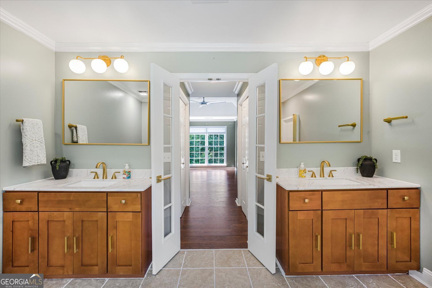 Bathroom with a double wood vanity, marble countertops, gold fixtures, and open double doors.