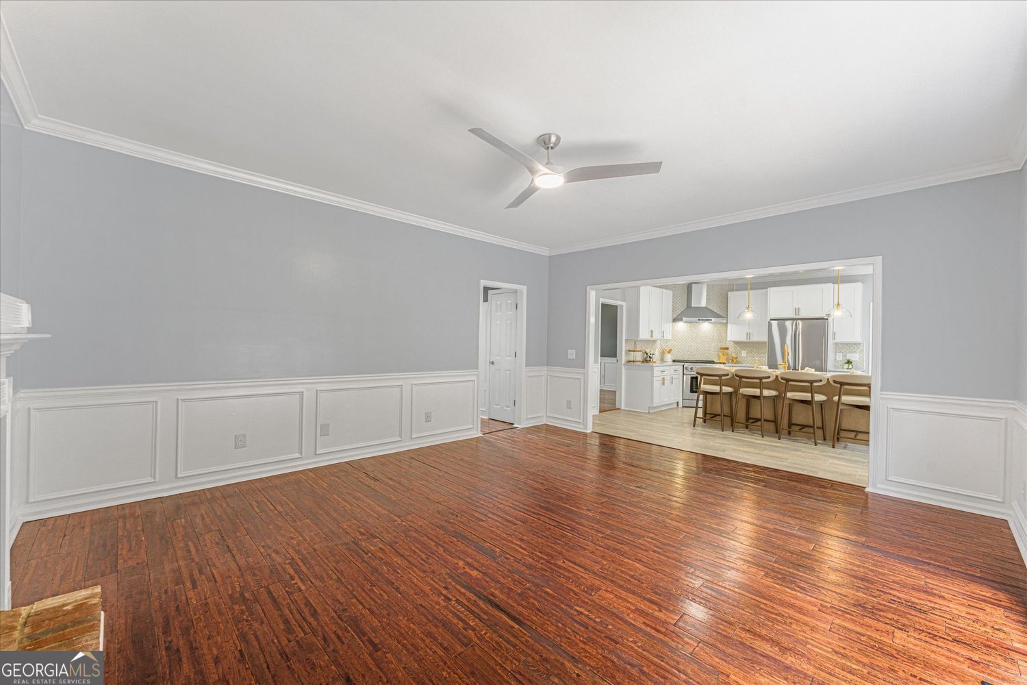 Empty room with polished hardwood floors and white wainscoting, opening into a bright kitchen.