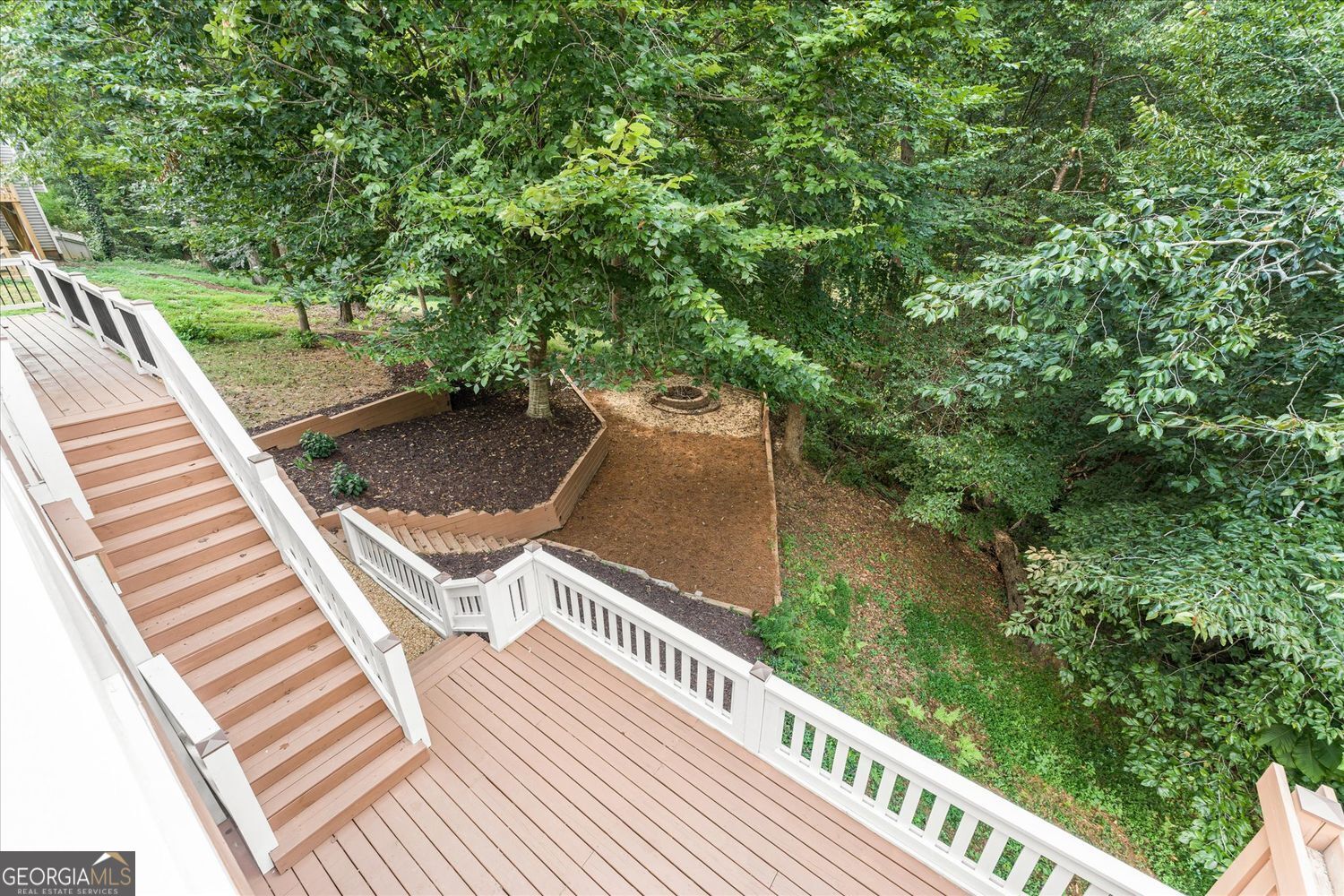 Wooden deck with white railings and stairs overlooking a wooded backyard with a fire pit.