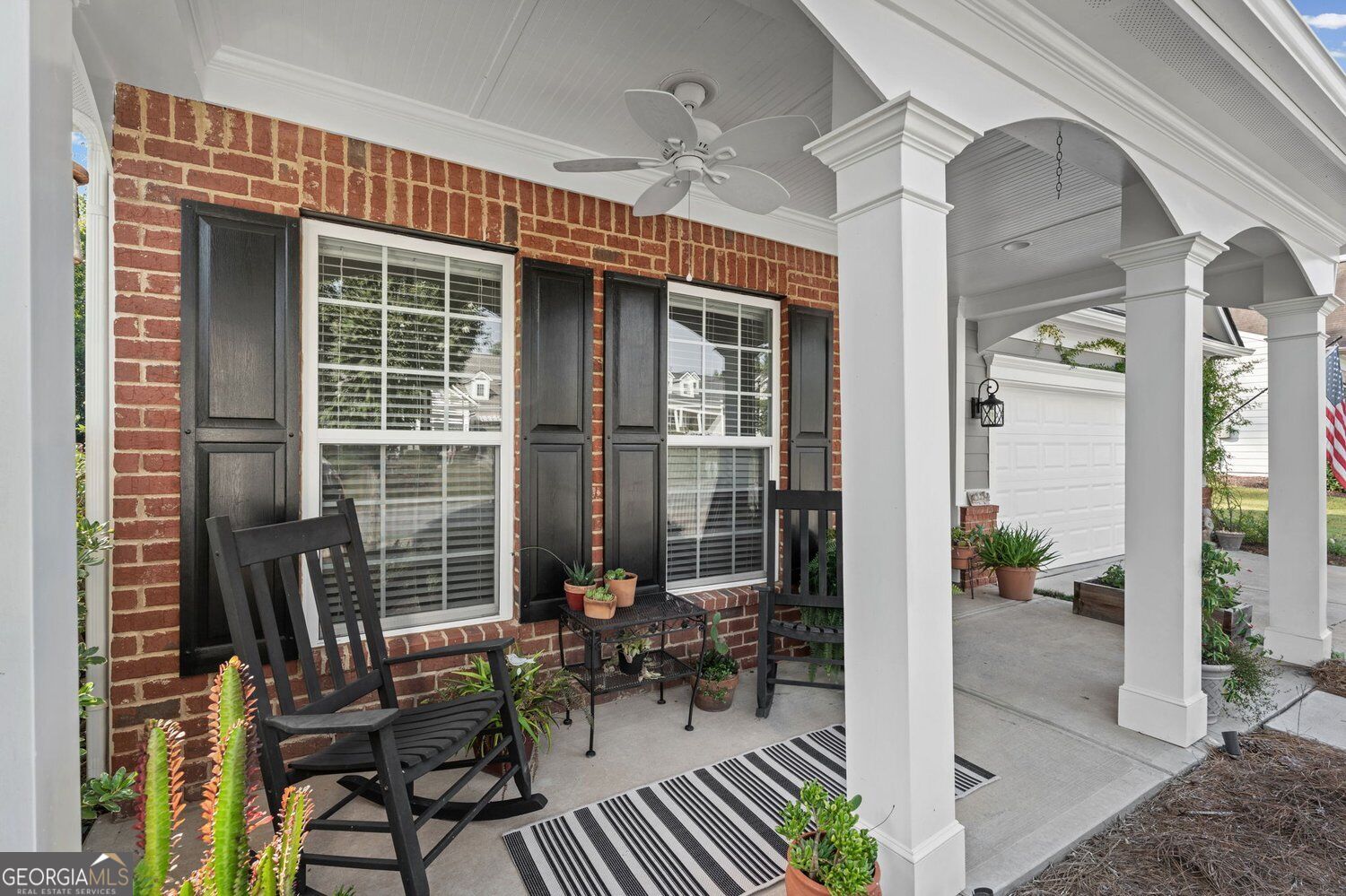 Brick home’s covered front porch with white arches, black rocking chairs, and potted plants.