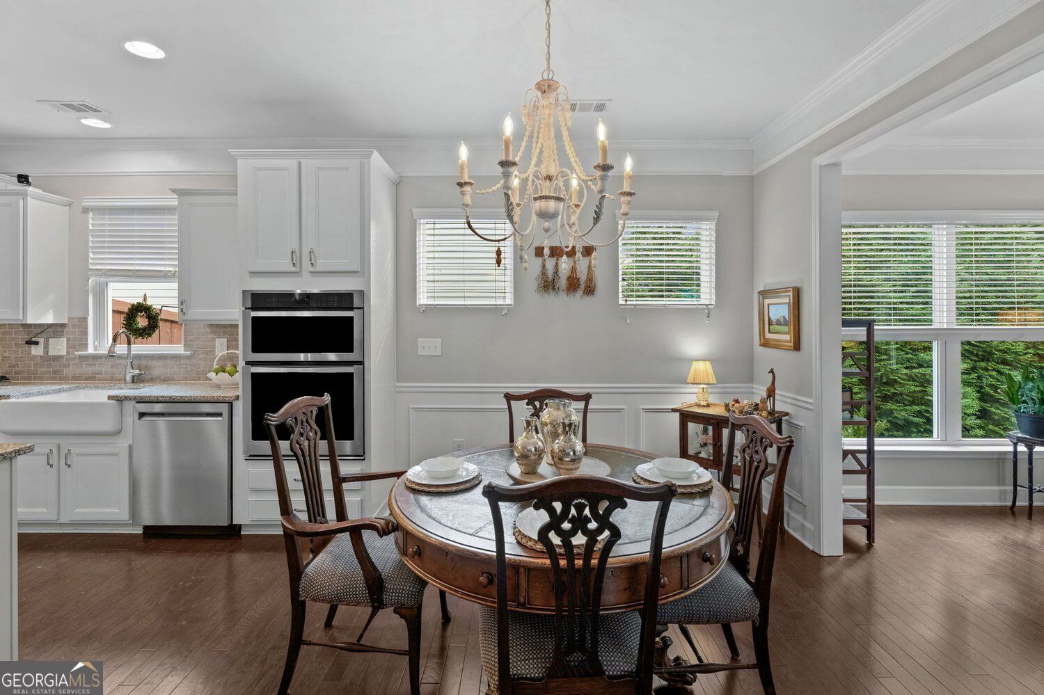 Dining room with a round wooden table, carved chairs, ornate chandelier, and adjoining white kitchen.