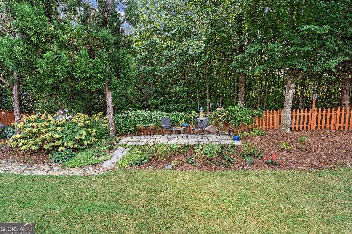 Backyard patio with outdoor seating, lush landscaping, mature trees, and a wooden picket fence.