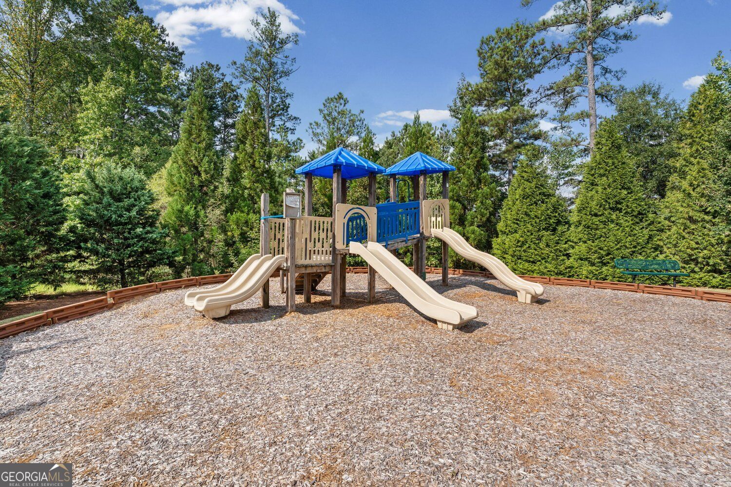 Community playground with a blue-roofed play structure, several slides, wood chips, and a green bench.