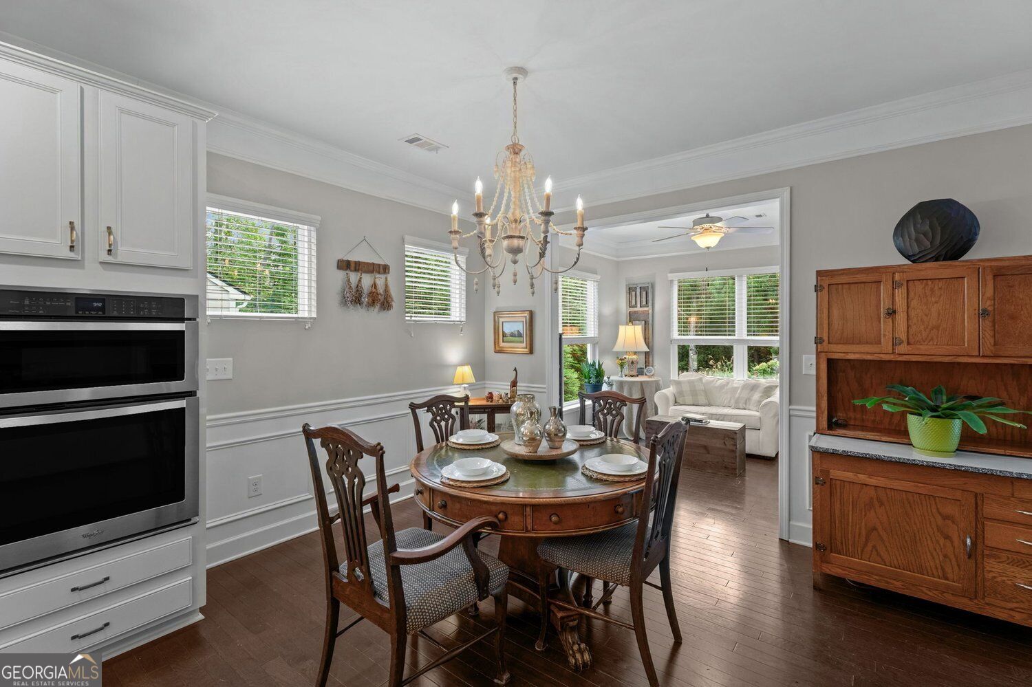 Dining room with a round wooden table beneath a chandelier, opening to a sitting room with a white sofa.