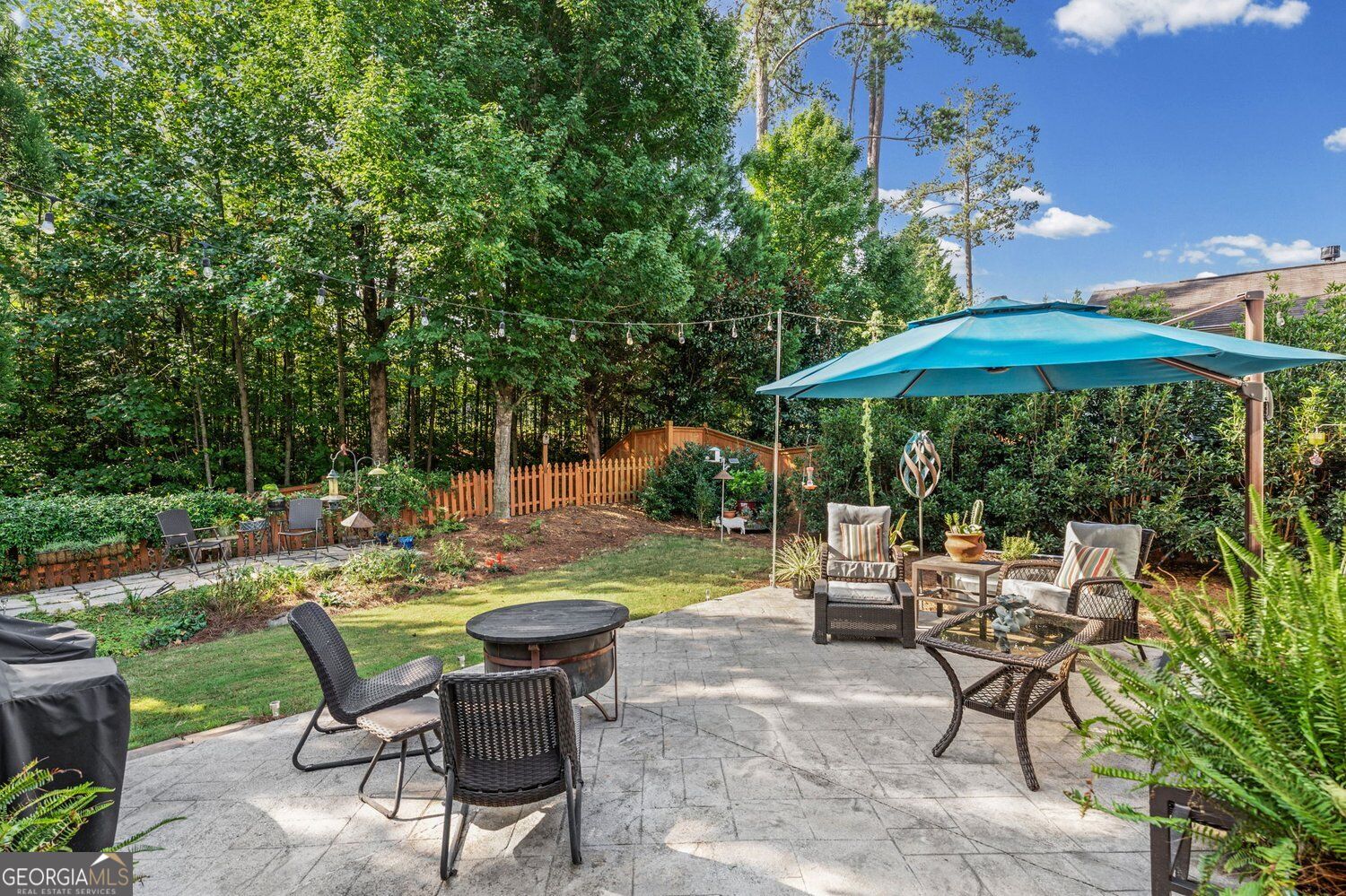 Backyard patio with wicker seating beneath a turquoise umbrella, beside a garden and wooded yard.