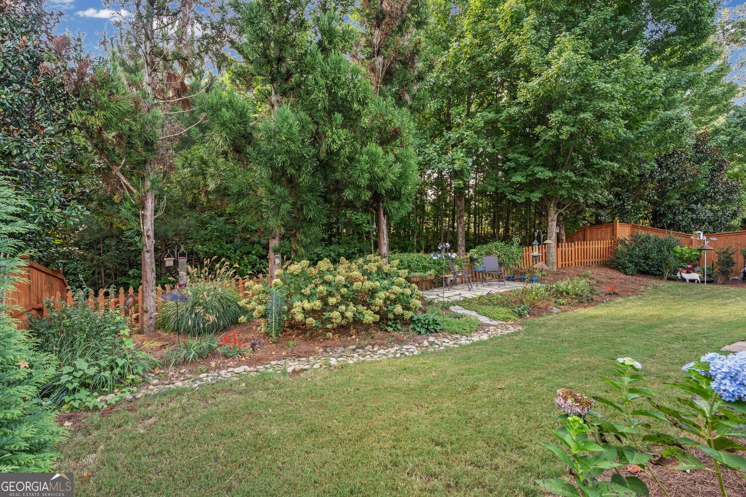 Landscaped backyard with mature trees, colorful garden beds, a lawn, and a small patio.