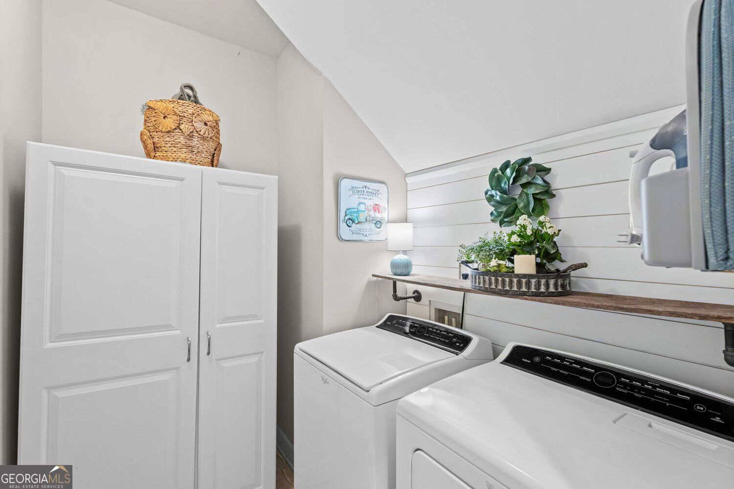 Laundry room with white washer and dryer, tall storage cabinet, and a decorative shelf against shiplap.