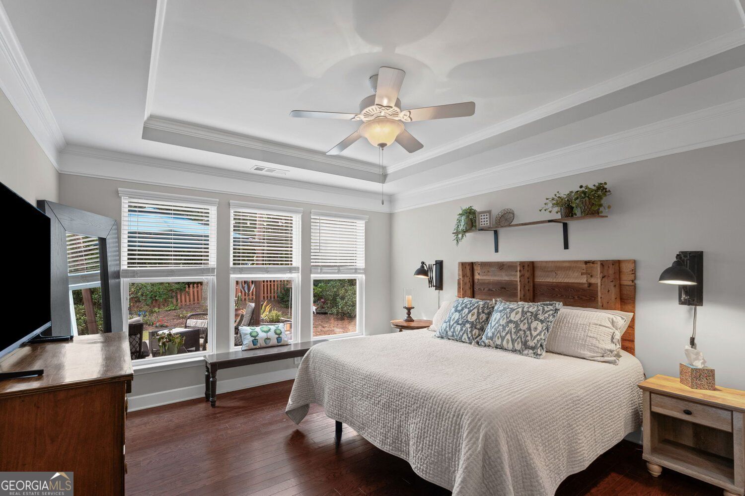 Bedroom with a rustic wood headboard, tray ceiling, ceiling fan, and windows overlooking a fenced garden.