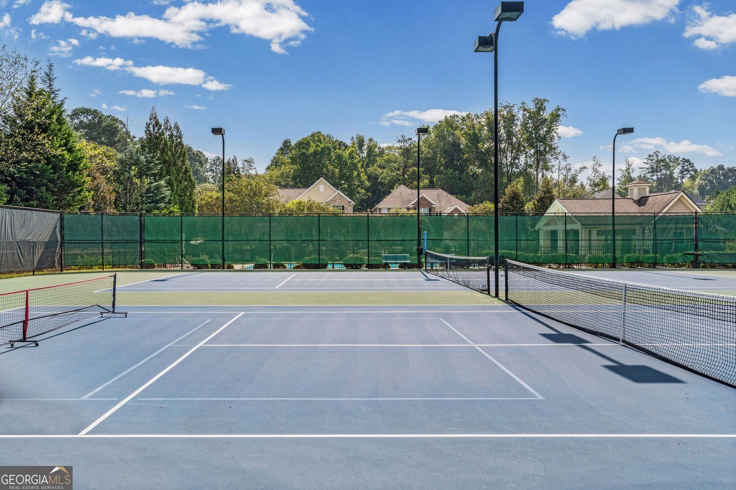 Outdoor tennis courts with green fencing, multiple nets, and tall lights.