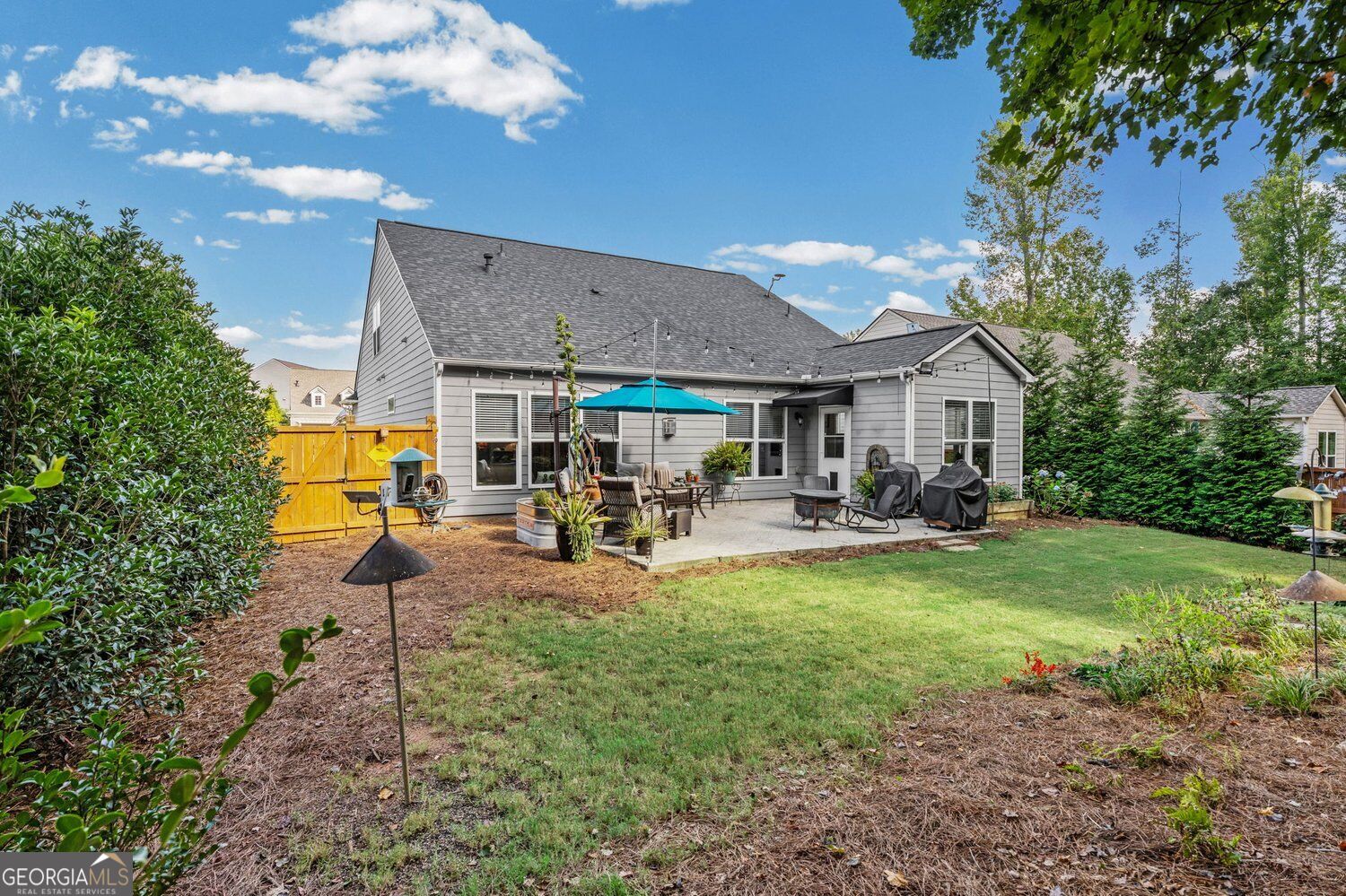 Gray home’s backyard patio with a turquoise umbrella, covered grills, yellow fence, and lawn.