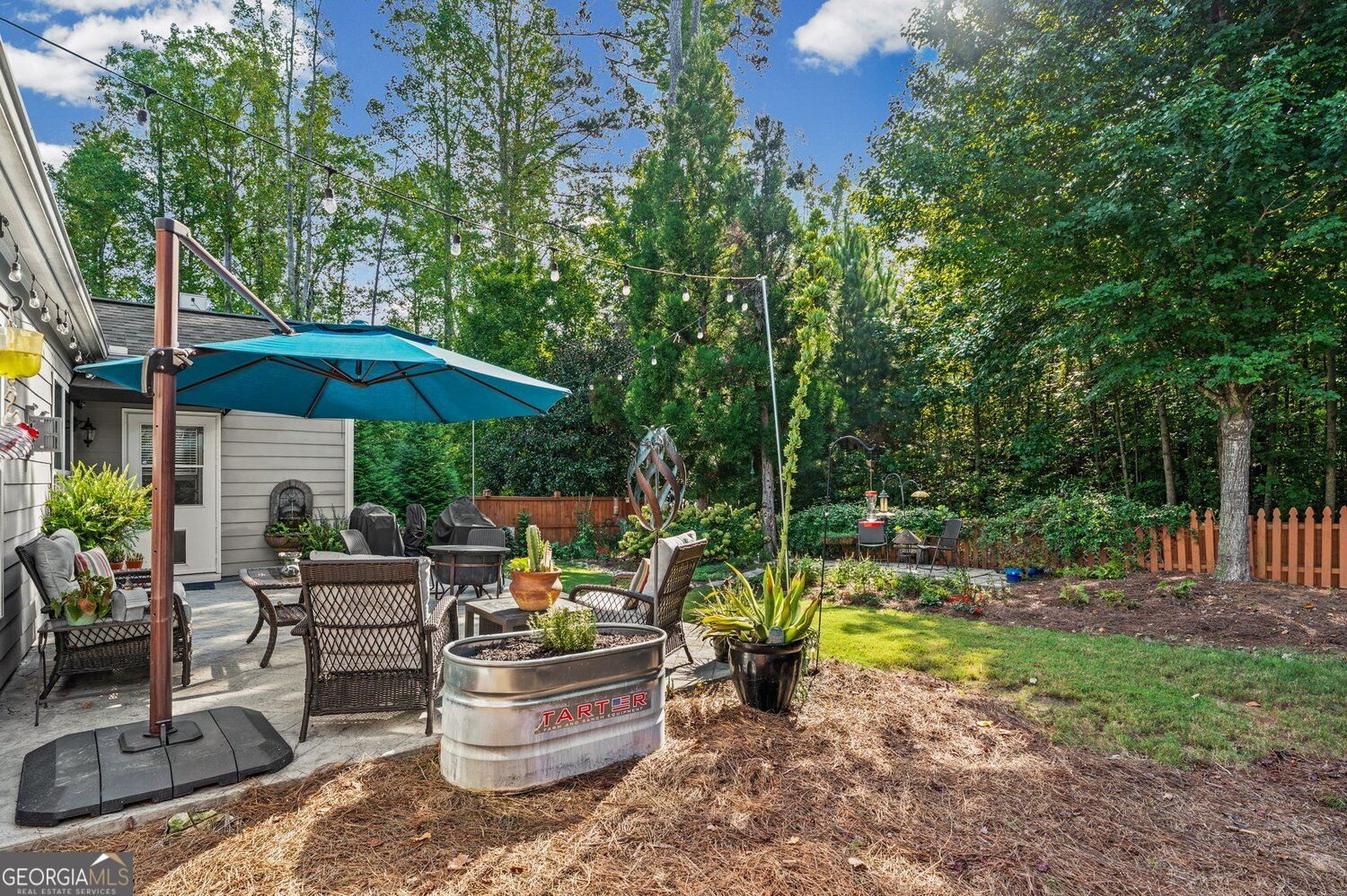 Backyard patio with wicker seating, turquoise umbrella, string lights, and mature trees.