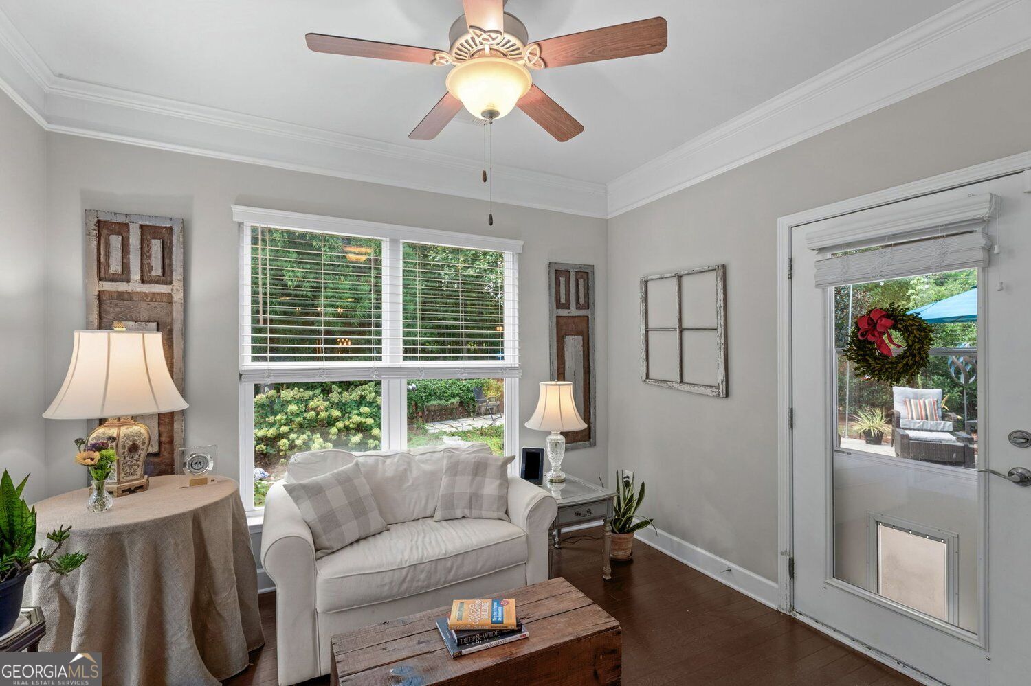 Furnished living room with a white loveseat, ceiling fan, and garden-facing windows beside a glass patio door.