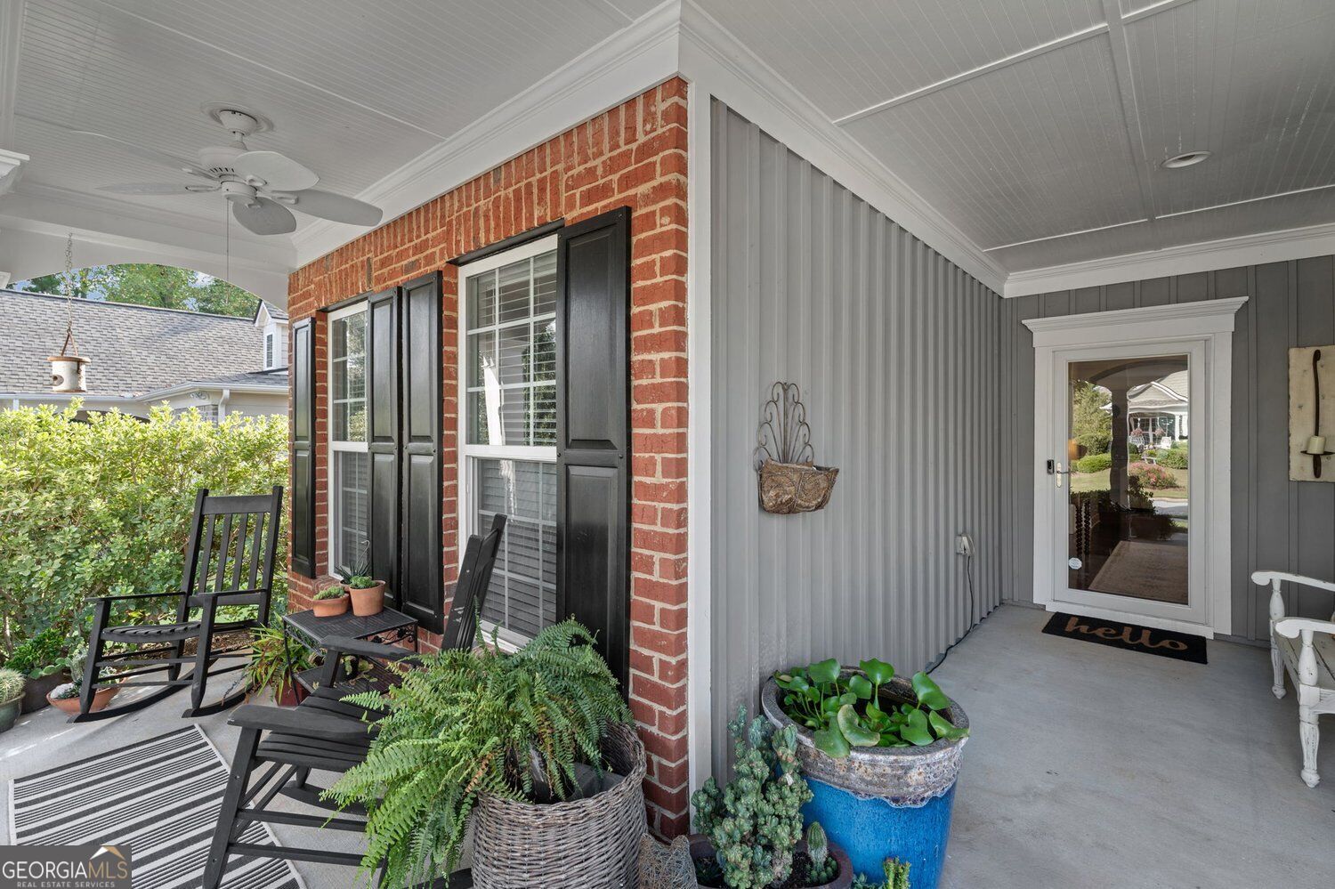 Covered front porch with brick walls, black rocking chairs, potted plants, and a glass entry door.