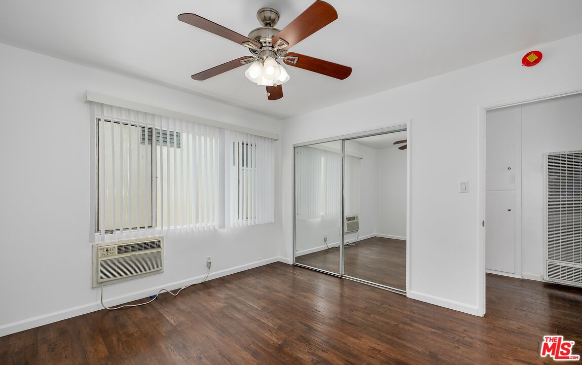 Empty apartment room with dark wood flooring, mirrored closet doors, window air conditioner, and ceiling fan.