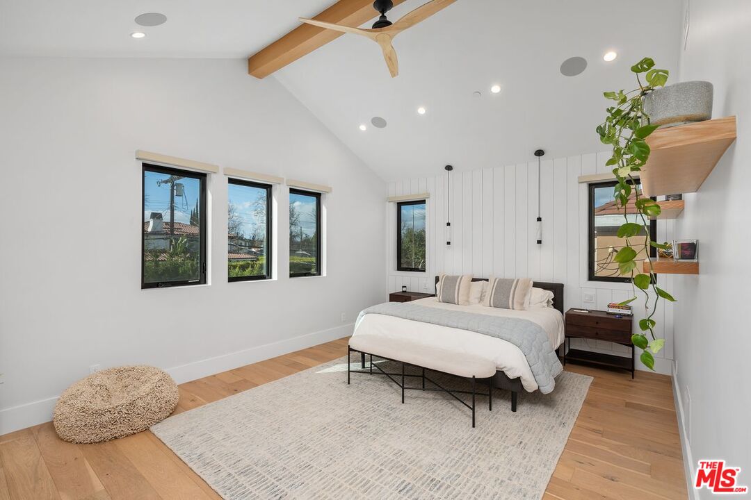 Staged bedroom with a white bed, paneled accent wall, exposed wood ceiling beams, and three windows.
