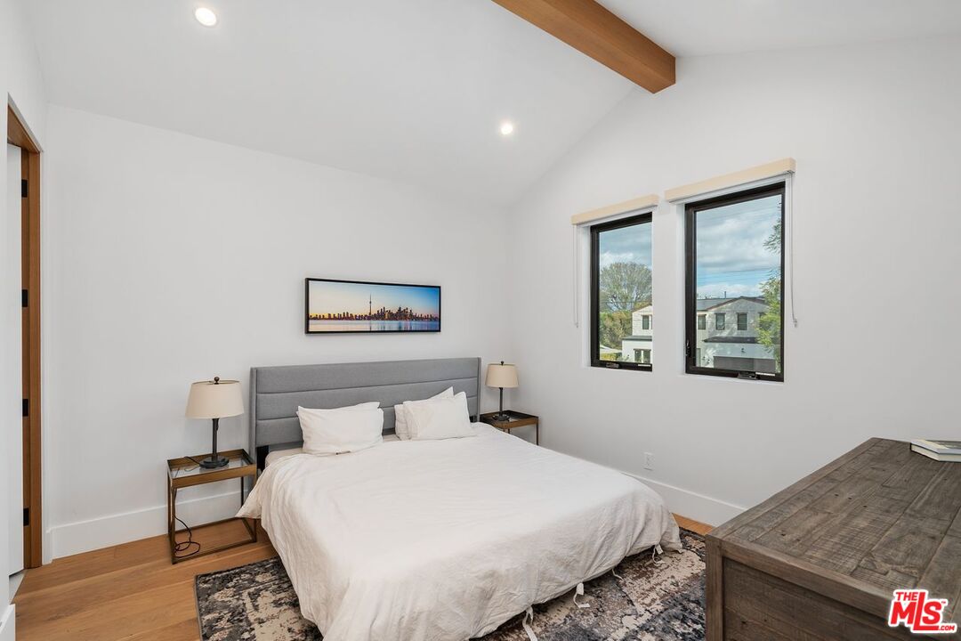 Furnished bedroom with a white bed, gray headboard, two windows, and an exposed wood ceiling beam.