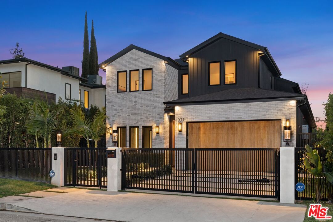Modern two-story home with white brick, dark siding, and a gated front yard at dusk.