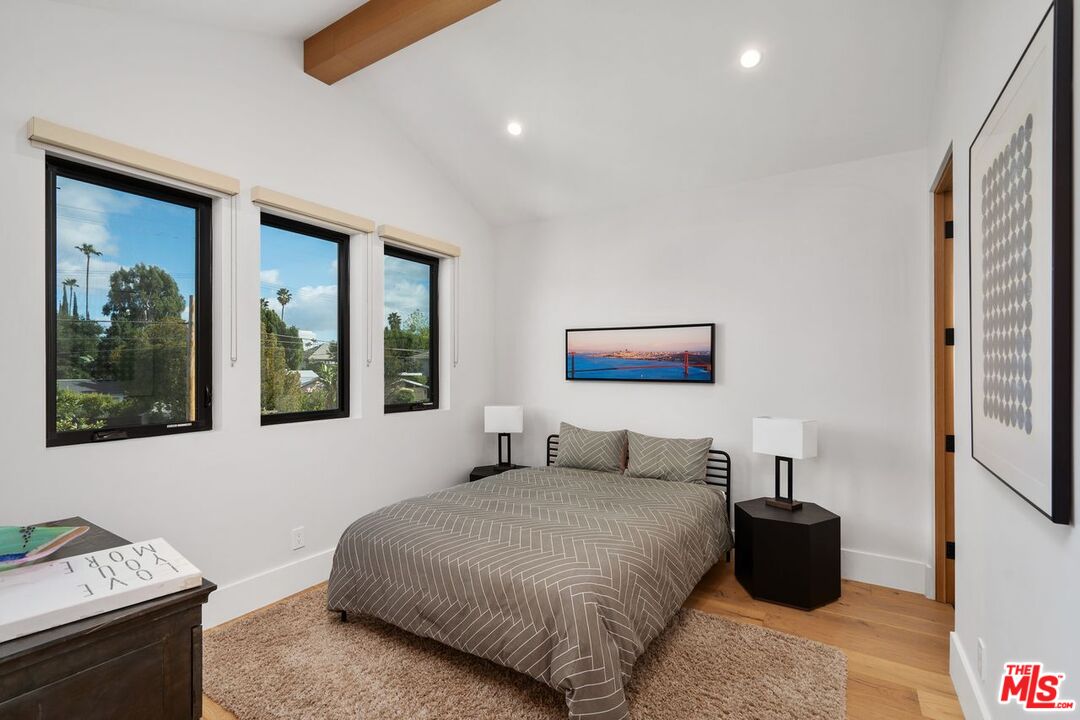 Furnished bedroom with a gray patterned bed, three tall windows, and a wood ceiling beam.