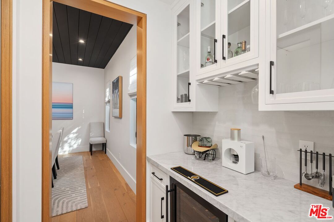 Wet bar with white glass-front cabinets, marble counters, and a doorway to a small seating area.